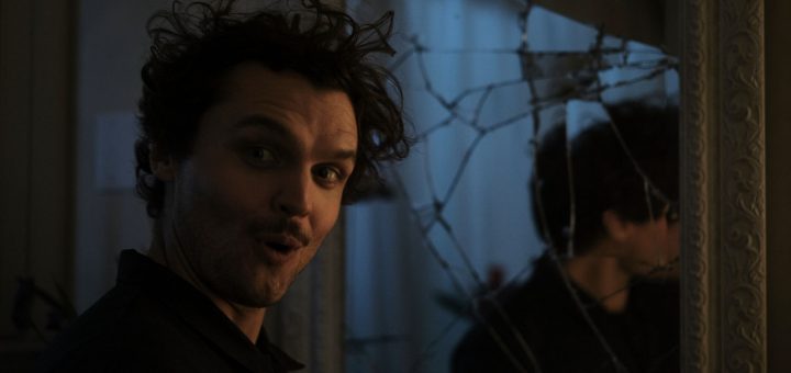 A person with curly hair and a surprised expression stands next to a large mirror with cracks, their reflection visible in the broken glass. The lighting is dim and moody.