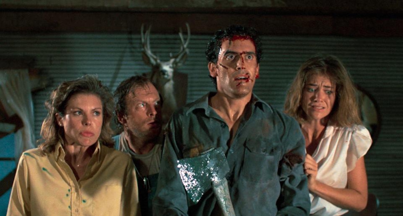 Four frightened people, one holding a bloody axe, stand close together in a dimly lit room with a deer head mounted on the wall behind them. They appear tense and distressed, with hints of blood on their faces and clothes.