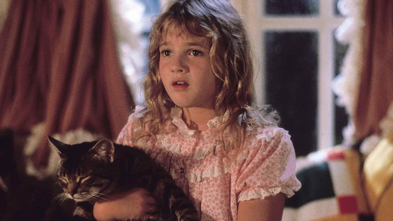 A young girl with wavy blond hair wearing a pink, ruffled nightgown holds a tabby cat. She looks ahead with a surprised expression. The background shows a curtained window and some cozy furnishings.