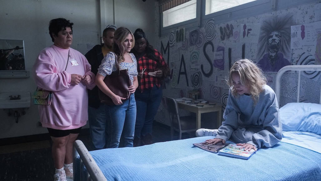 Five people stand in a dimly lit, graffiti-covered room, watching a woman in gray sit on a bed and look at a book. The tense atmosphere suggests these could be cursed friends facing an unsettling mystery together.