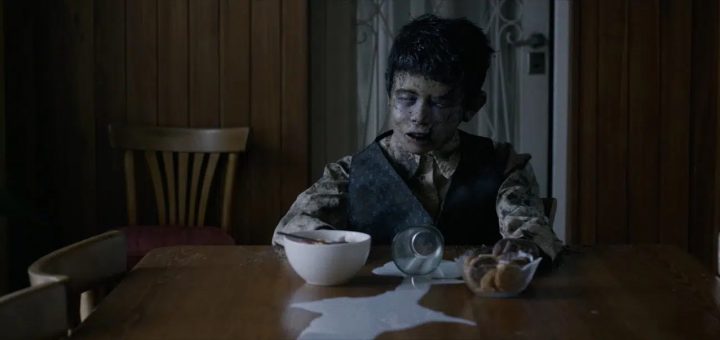 A boy with darkened skin and hollow eyes sits at a wooden table, staring ahead blankly. A spilled glass of milk, a bowl, and a dish of cookies are in front of him. The room has wooden walls and an empty chair beside him.