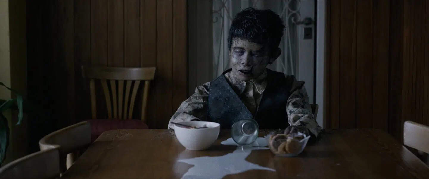 A boy with darkened skin and hollow eyes sits at a wooden table, staring ahead blankly. A spilled glass of milk, a bowl, and a dish of cookies are in front of him. The room has wooden walls and an empty chair beside him.