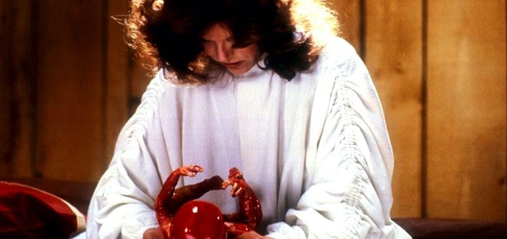 A woman in a white robe with wavy brown hair looks down at and holds a small, red, unnatural-looking creature with tiny arms. The background is wood-paneled.
