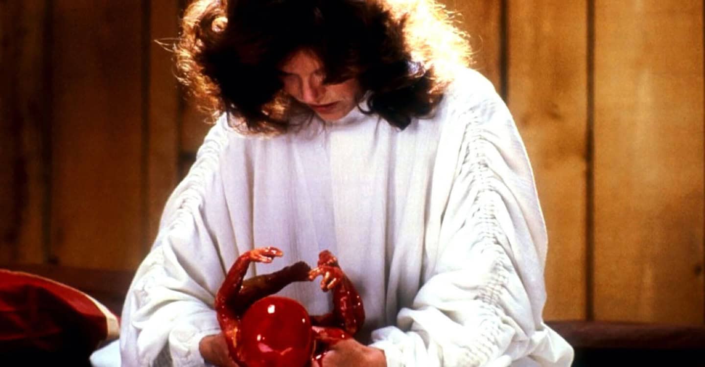 A woman in a white robe with wavy brown hair looks down at and holds a small, red, unnatural-looking creature with tiny arms. The background is wood-paneled.