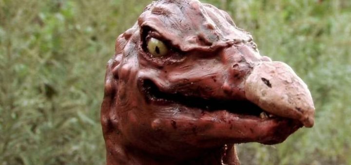 A close-up of a Thankskilling creature with reddish-brown, wrinkled skin, a large beak-like nose, sharp teeth, and yellow reptilian eyes, set against a blurred green outdoor background.