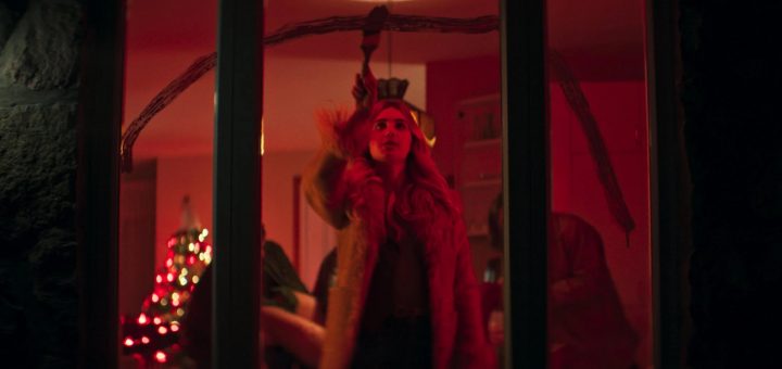 A person with long hair wearing a fur coat stands inside a warmly lit room, reaching up toward a branch hanging from the ceiling. In the background, a decorated Christmas tree glows amid festive, reddish lighting—evoking scenes from a classic Christmas movie.