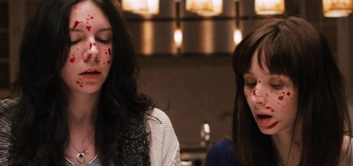 Two women with long dark hair and pale skin stand indoors with blood splattered on their faces. Both have their eyes closed or looking down, appearing distressed. Warm pendant lights hang in the blurred background.