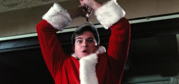 A person dressed in a red Santa Claus costume holds an object overhead with both hands, looking intense and startled—evoking a scene straight out of Silent Night Deadly Night 2. The background shows part of a ceiling and a doorway.