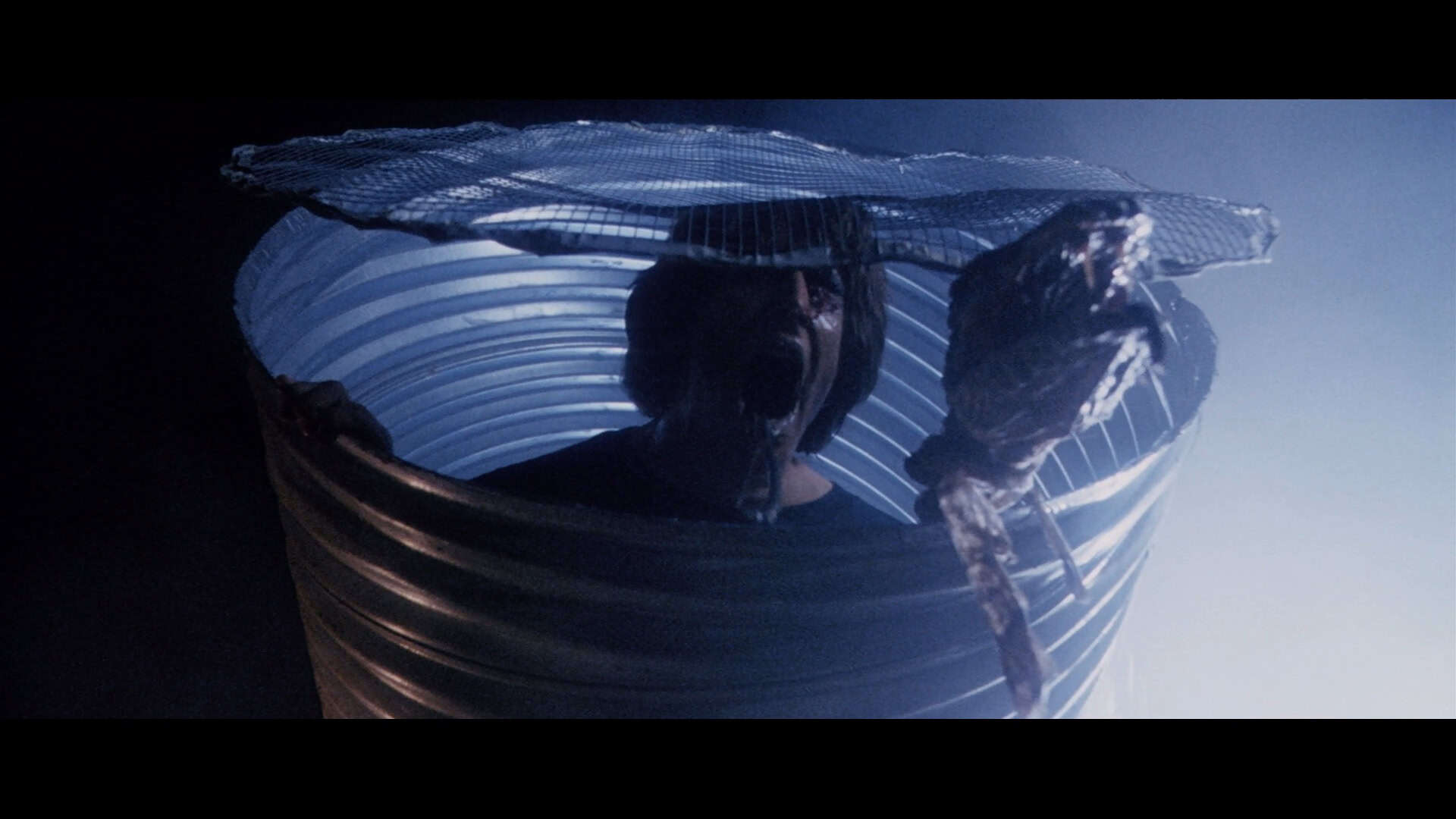 A person emerges from a large metal barrel with a mesh cover, holding up what appears to be a severed, bloody arm in a dark, eerie setting with blue lighting.
