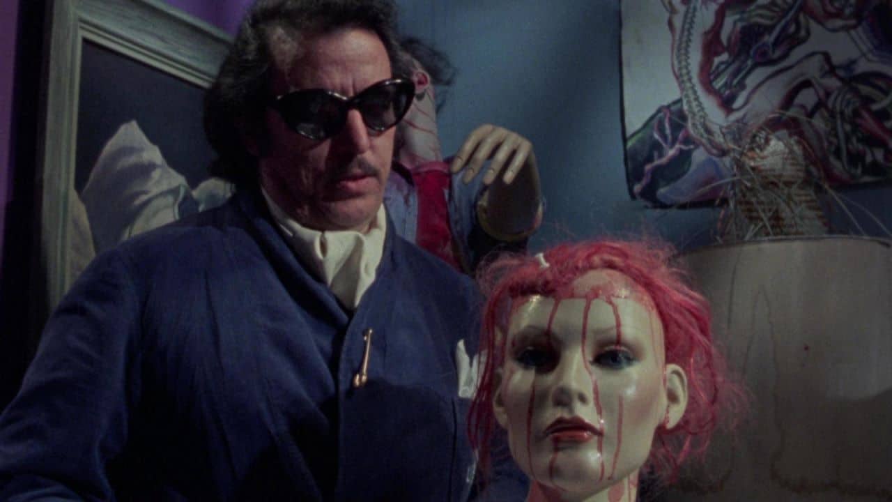 A man in dark sunglasses and a blue suit stands behind a mannequin head with red hair and fake blood running down its face, in a dimly lit, cluttered room with eclectic art and mannequin parts.