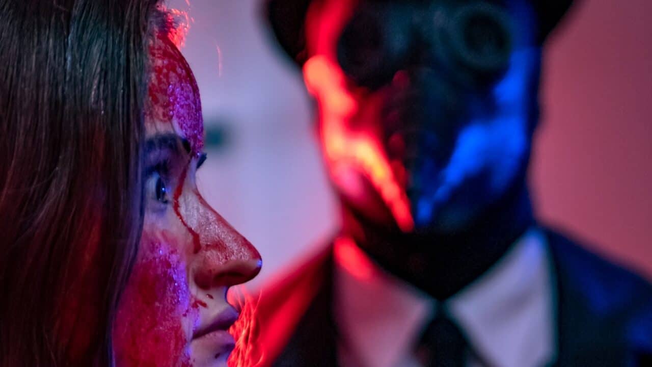 A close-up of a woman with a bloodied face looking nervously to the side, while a masked figure with a beak-like nose stands ominously in the background under red and blue lighting—setting an eerie tone for New Fears Eve.