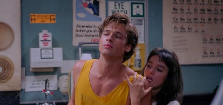A young man in a yellow tank top stands with a relaxed expression as a young woman leans on his shoulder, holding his arm. Behind them are wall posters, safety signs, and an eye wash station in a classroom or lab setting.
