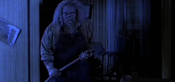 A disheveled person with long, wild hair and a menacing expression holds an axe inside a dimly lit room with blue lighting, creating a tense and eerie atmosphere.
