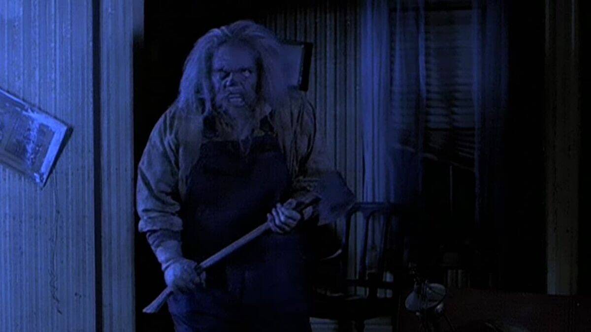 A disheveled person with long, wild hair and a menacing expression holds an axe inside a dimly lit room with blue lighting, creating a tense and eerie atmosphere.