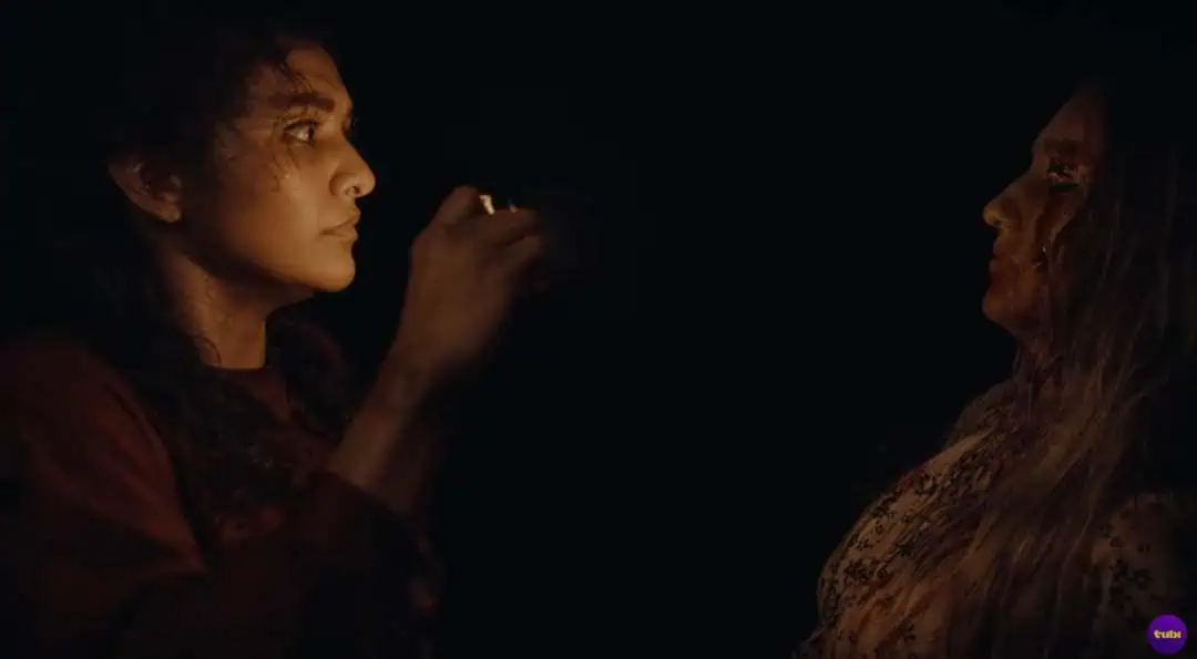 Two women face each other in a dark setting; one holds a lighter up, illuminating both their faces. The other woman has long hair and blood on her face, creating a tense atmosphere.