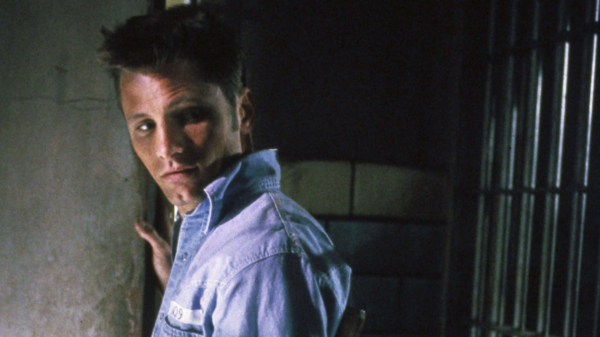 A man with short hair in a light blue prison uniform stands near a wall, looking over his shoulder toward the camera. Behind him, metal prison bars are visible, suggesting a jail setting.