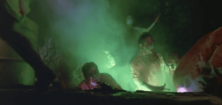 Several people with bloodied faces emerge from a dark pit filled with glowing green smoke, creating a dramatic and eerie atmosphere.