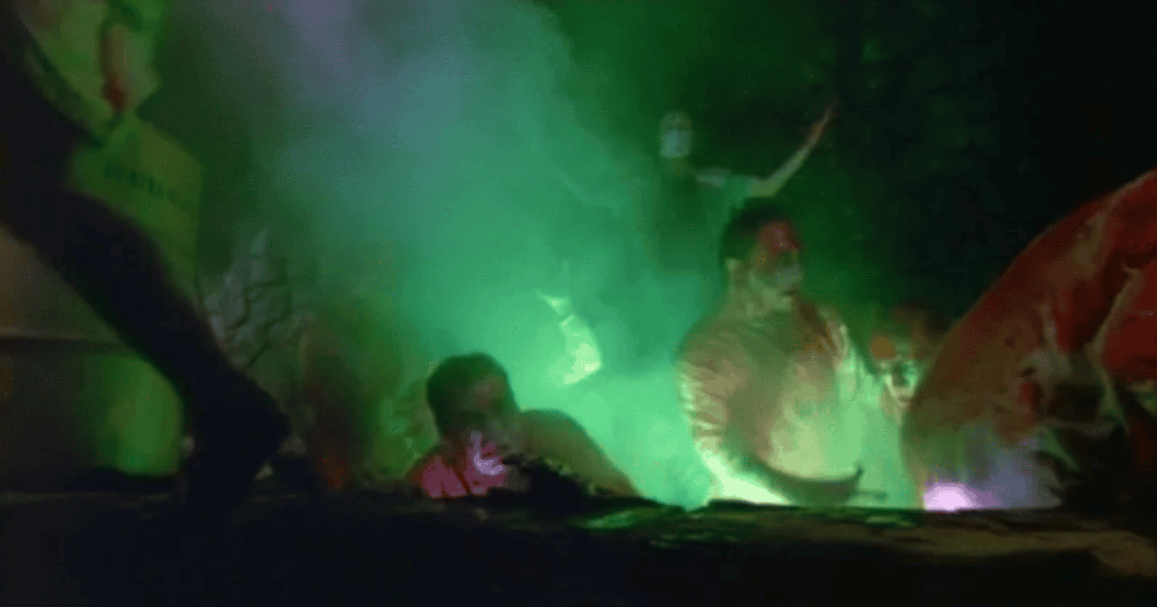 Several people with bloodied faces emerge from a dark pit filled with glowing green smoke, creating a dramatic and eerie atmosphere.