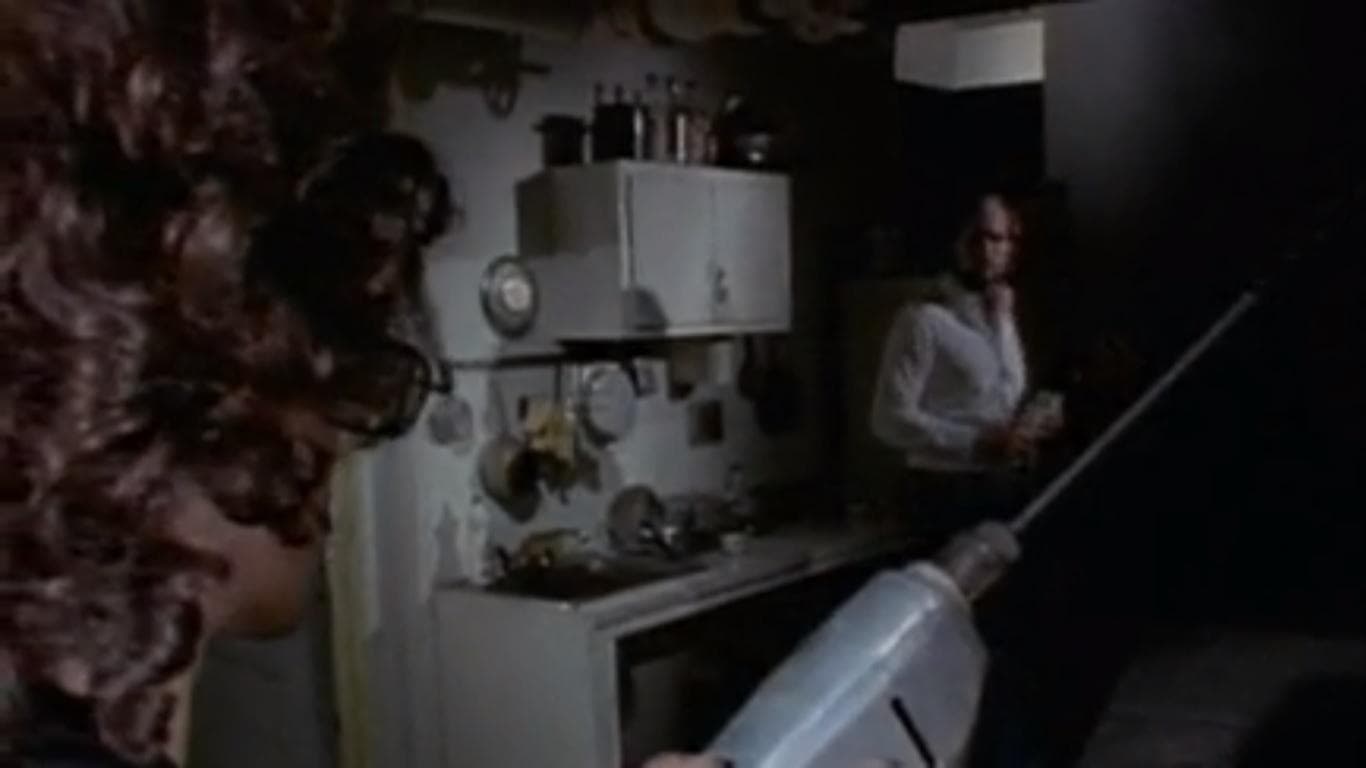 A person with curly hair holds a large electric drill in the foreground, aiming at another person standing in a dimly lit kitchen area cluttered with dishes and bottles.