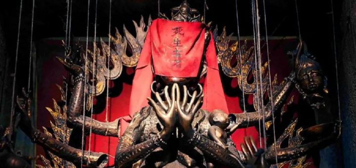 A multi-armed statue with a red cloth covering its face, surrounded by intricate golden details and additional heads, stands against a red background. The hands are raised in various gestures.