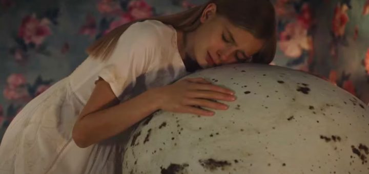A young woman in a white dress leans her head and arms tenderly against a large, speckled egg. The background is decorated with pink floral patterns.