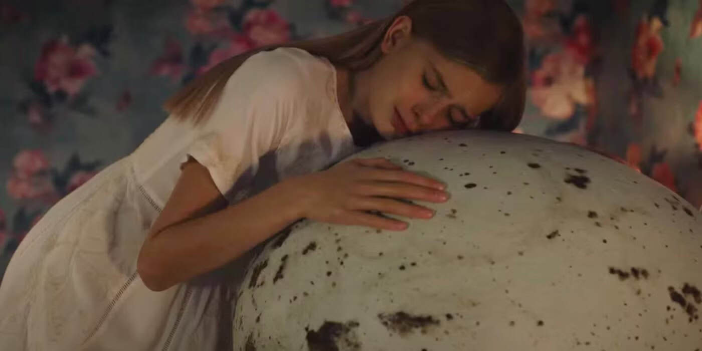 A young woman in a white dress leans her head and arms tenderly against a large, speckled egg. The background is decorated with pink floral patterns.