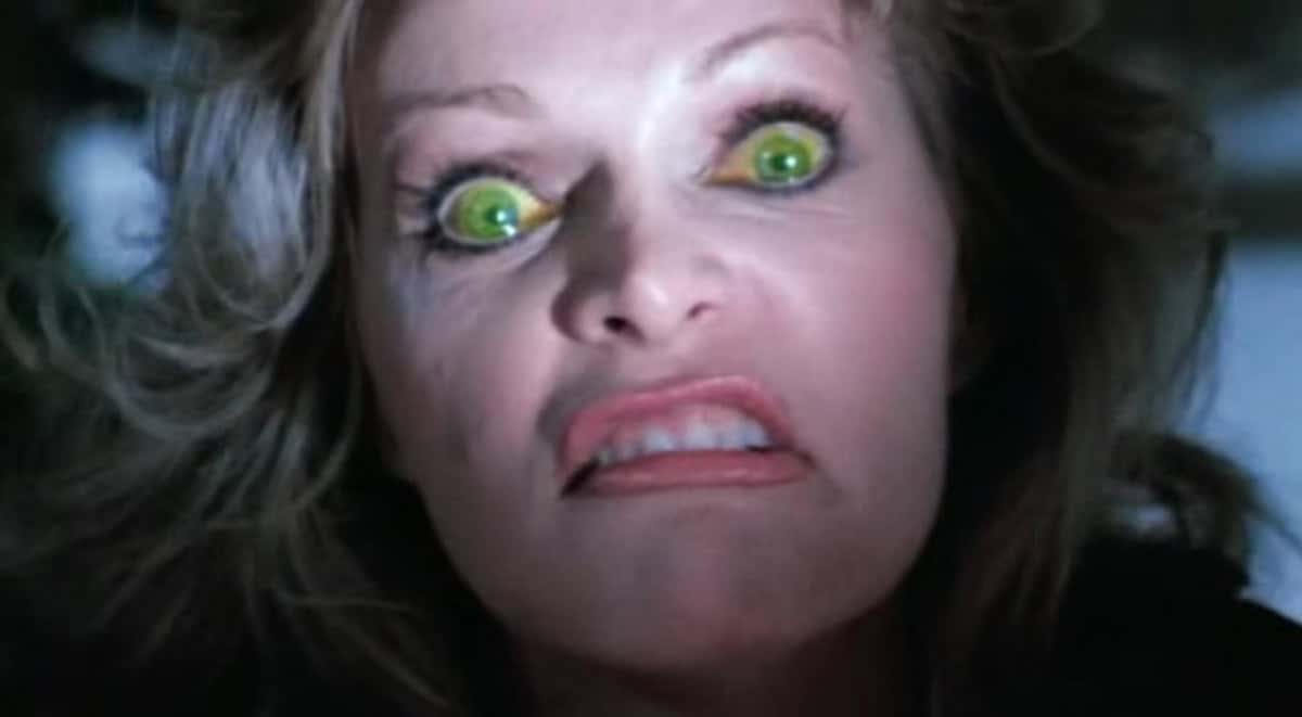 A woman with wide, green glowing eyes and a contorted, angry expression stares intensely at the camera, her teeth clenched and lips curled back—certainly not the usual "Happy Birthday To Me" mood.