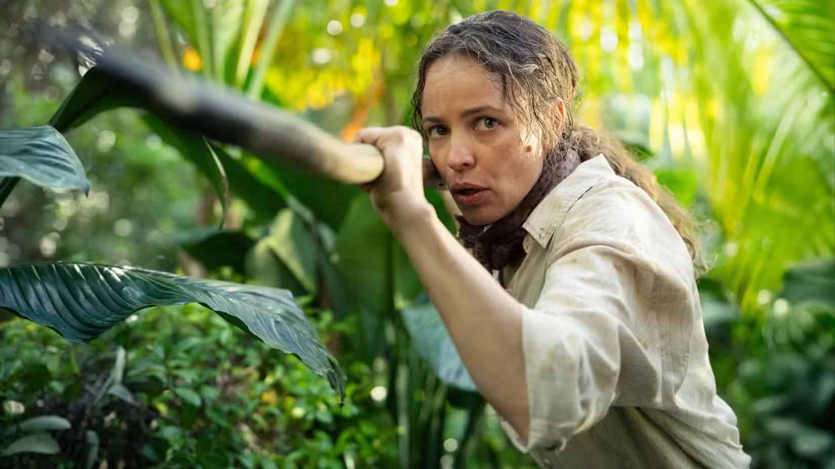 A woman in a light shirt crouches in dense green jungle foliage, clutching a long spear with a determined expression—ready for an adventurous birthday celebration as sunlight filters through the leaves.