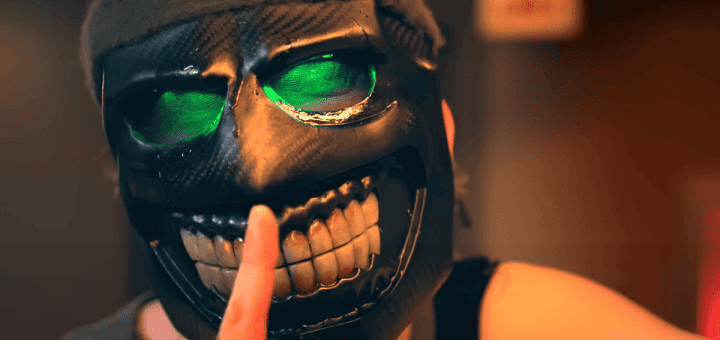 A person wearing a black, grinning mask with glowing green eyes holds a finger to their lips in a "shh" gesture, signaling silence. The background is blurred.