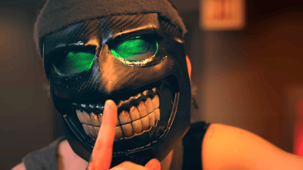 A person wearing a black, grinning mask with glowing green eyes holds a finger to their lips in a "shh" gesture, signaling silence. The background is blurred.