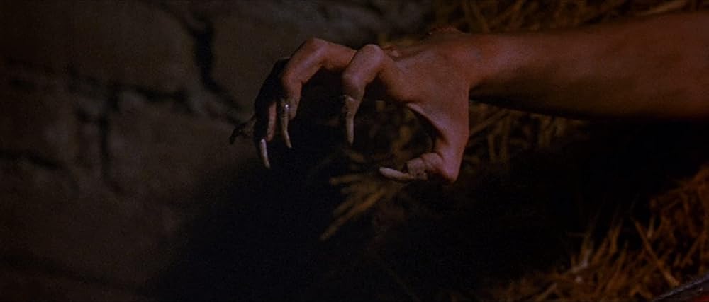 A close-up of a hand transforming into a clawed, monstrous shape with long, pointed fingernails, set against a dimly lit, rough background with a hint of straw.