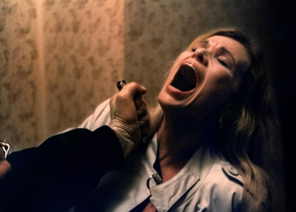 A woman with blonde hair screams in fear, her mouth wide open, as a hand holding a knife approaches her throat in a dimly lit, tense scene reminiscent of the terror in New York Ripper.