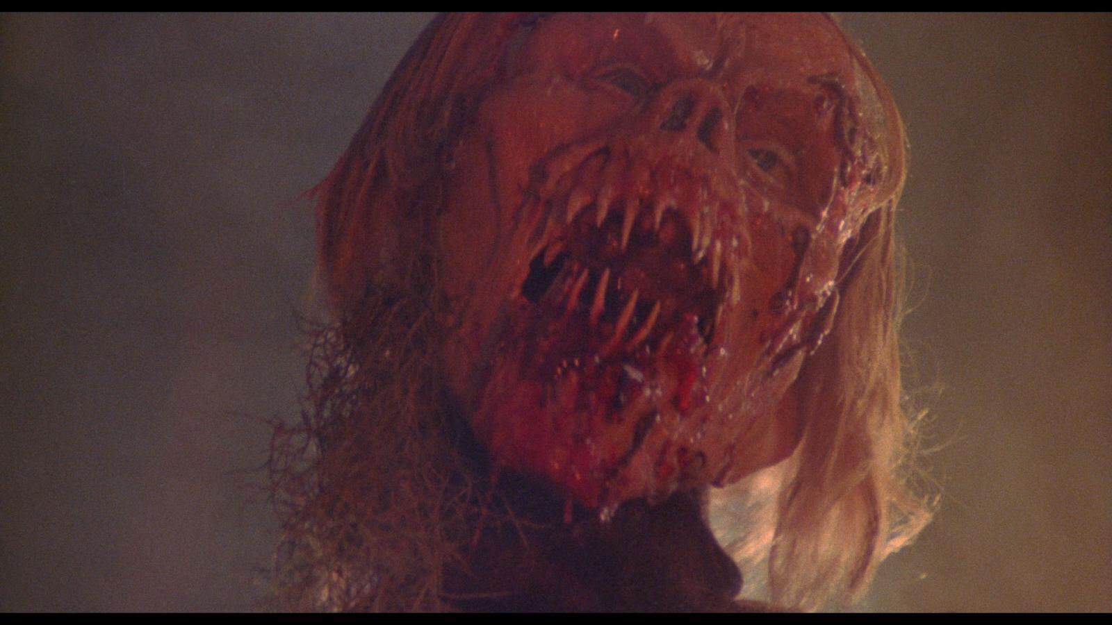 A gruesome, partially decomposed face with long blond hair, exposed bone, and blood-covered mouth forms a horrifying scene—perfect for a twisted Happy Birthday scare in dim lighting.