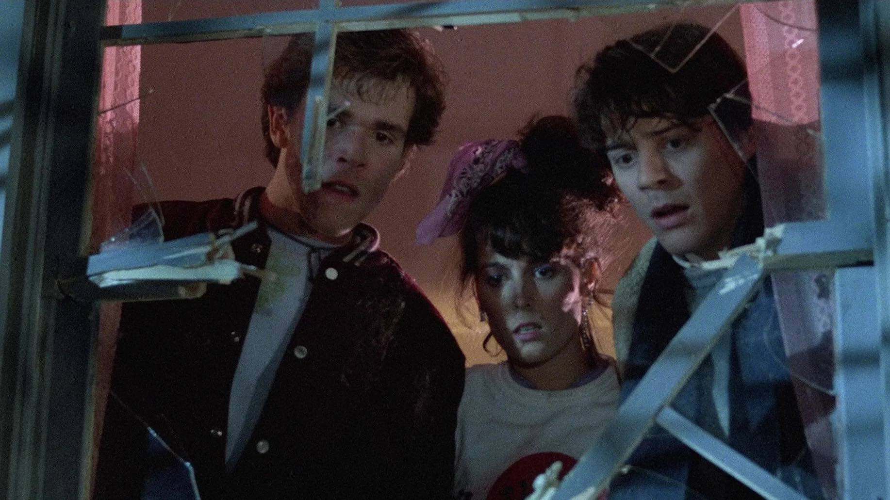 Three young people, two males and one female with a pink scrunchie, look down in shock through a jagged broken window, dim light casting eerie shadows. The suspenseful scene evokes the unsettling atmosphere of Color Out Of Space.
