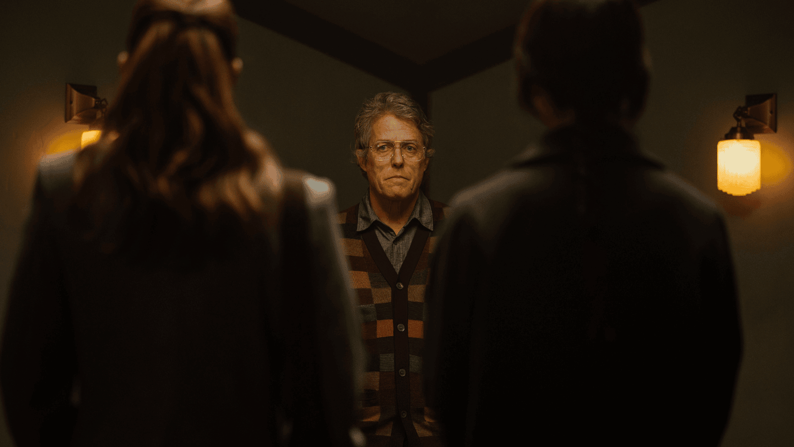 An older man in glasses and a colorful striped cardigan stands facing two people whose backs are to the camera, in a dimly lit room with wall sconces—an atmosphere reminiscent of Color Out Of Space.