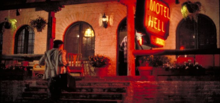 A man carrying a suitcase walks up the steps toward the entrance of a building with a glowing red neon sign that reads "Motel Hell" at night. The scene is dimly lit, evoking the eerie atmosphere of Color Out Of Space.