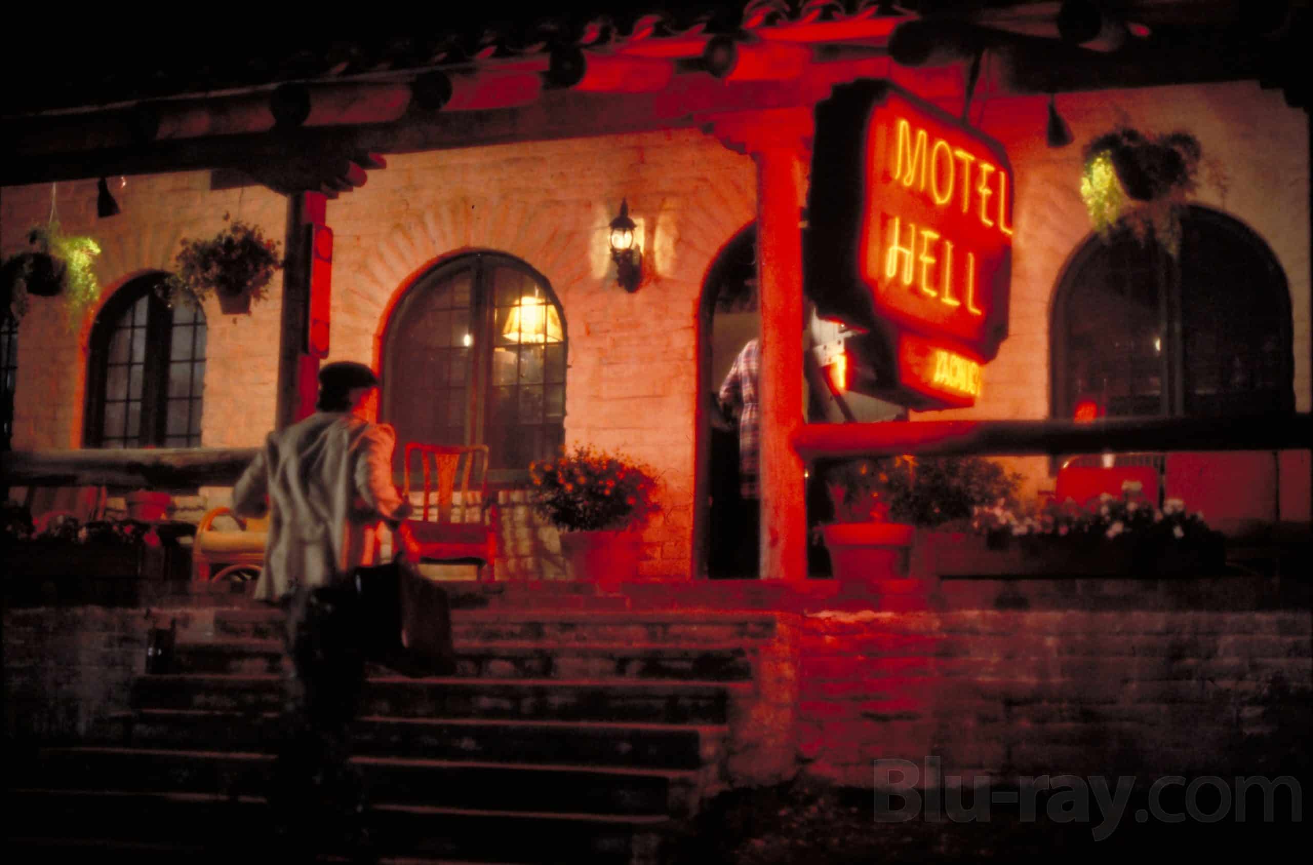 A man carrying a suitcase walks up the steps toward the entrance of a building with a glowing red neon sign that reads "Motel Hell" at night. The scene is dimly lit, evoking the eerie atmosphere of Color Out Of Space.
