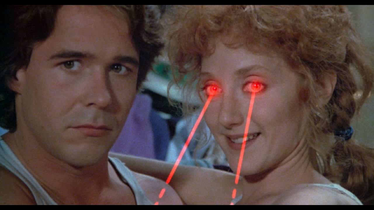 A man and woman sit close together. Channeling a sci-fi horror vibe inspired by H.P. Lovecraft’s Color Out Of Space, the woman smiles as bright red laser beams shoot from her eyes, while the man looks ahead with a neutral expression.