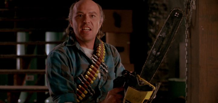 A man in a plaid shirt with a shotgun shell bandolier holds a large chainsaw indoors, looking intensely forward. Cardboard boxes and stairs are visible in the background.